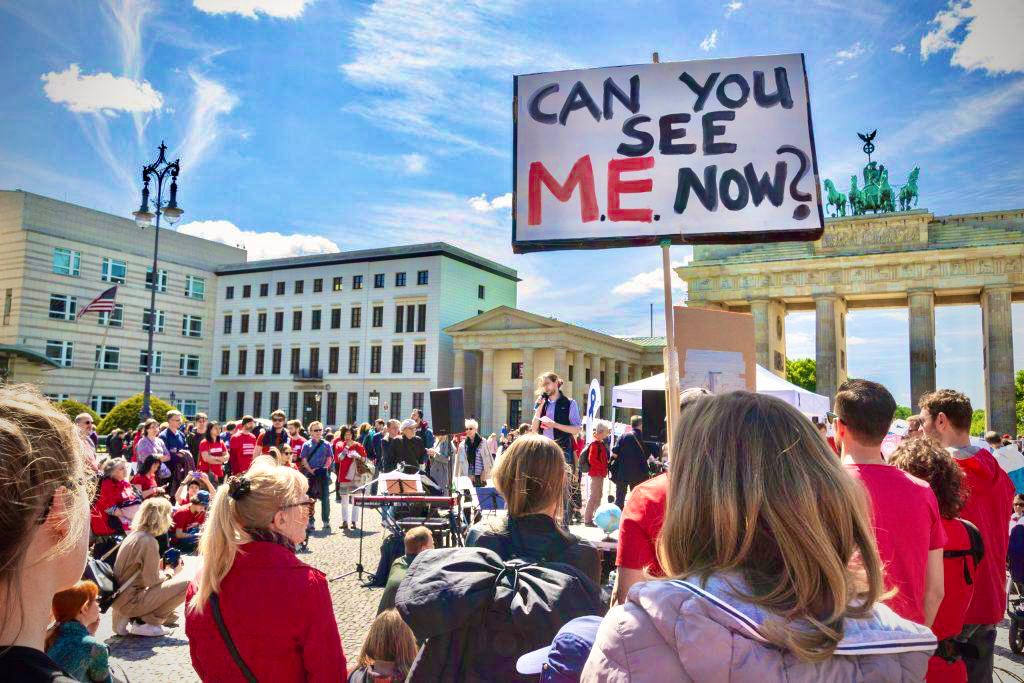 Aktion vor dem Brandenburger Tor mit mehreren Menschen, jemand hält ein Plakat mit dem Text: Can you see M.E. now?